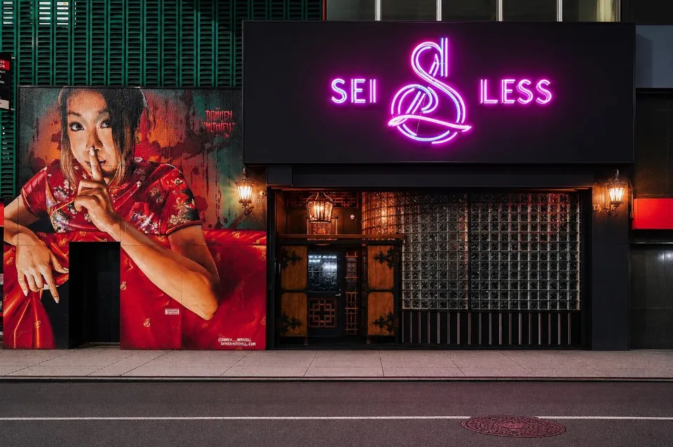 Sei Less storefront at 156 West 38th Street in Midtown Manhattan — pink neon sign above the entrance and the Damien Mitchell mural